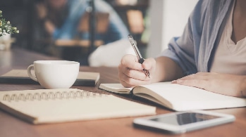 Student writing notes at desk with cup and smartphone.